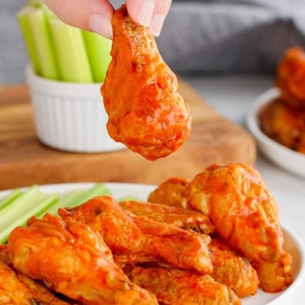 A hand holds up a buffalo wing over a plate of more wings and celery sticks.